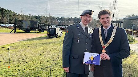 De commandant en de burgemeester staan buiten ter gelegenheid van het 150-jarig bestaan van de Verbindingstroepen. Op de achtergrond staan militaire voertuigen.